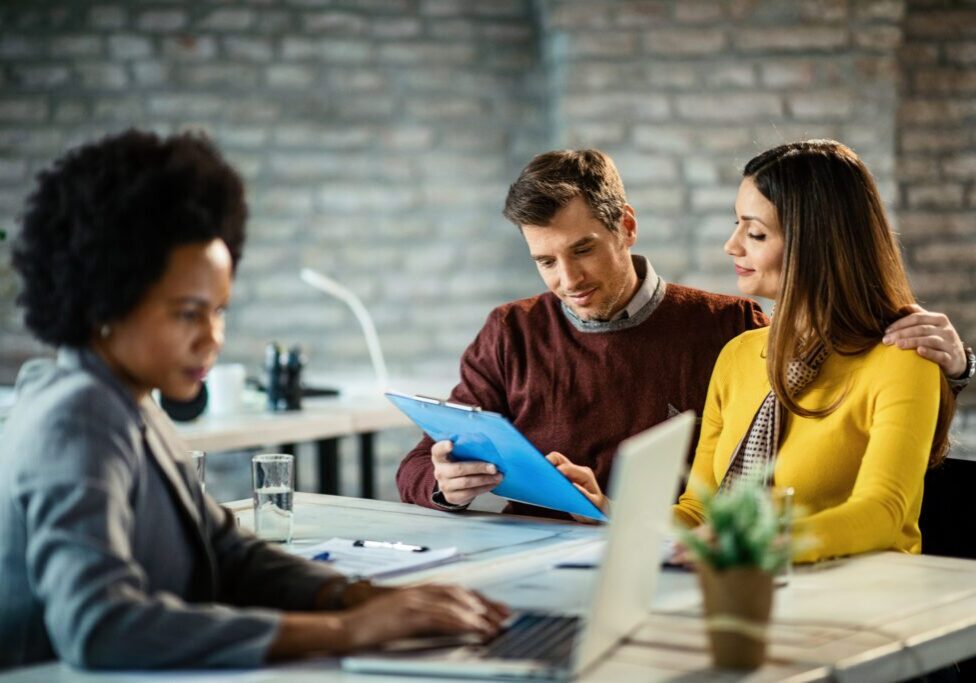 Smiling couple analyzing their financial reports on a meeting with insurance agent. African American woman at desk with white couple